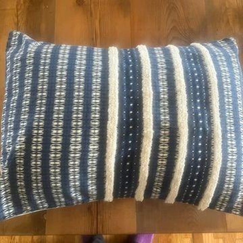 May include: Rectangular decorative pillow with a navy blue and white striped pattern. The pillow features vertical stripes of navy blue fabric with a textured white stripe in between. The pillow is on a wooden surface.