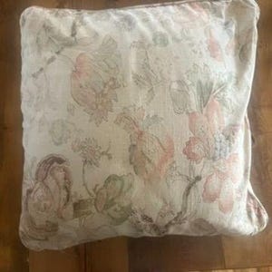 May include: Square decorative pillow with a floral pattern in shades of pink, green, and beige. The pillow has a soft, textured appearance and is likely made of fabric. The floral design features large blooms and foliage.