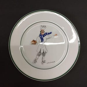 May include: White ceramic plate with a green rim and a central illustration of a chef figure. The chef is wearing a white uniform, a blue and white scarf, and is holding a pie. The plate has a glossy finish.