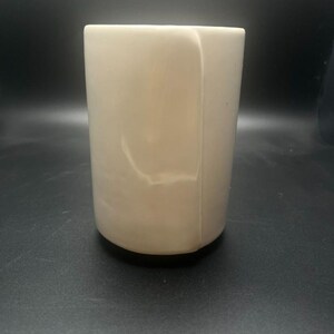 May include: A cylindrical, off-white tumbler with a rounded base. The smooth surface has subtle marbling and a slight sheen. The cup is empty and appears to be made of a solid material.