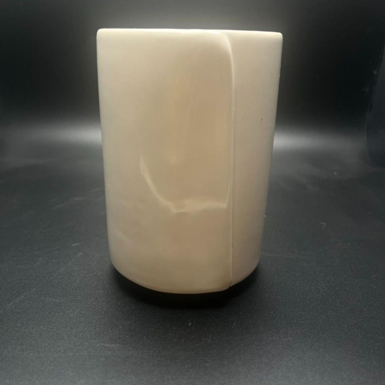 May include: A cylindrical, off-white tumbler with a rounded base. The smooth surface has subtle marbling and a slight sheen. The cup is empty and appears to be made of a solid material.