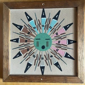 May include: A framed sand painting featuring a stylized sun design. The sun has a green face with black eyes and mouth, surrounded by radiating elements in black, blue, pink, and brown. The artwork is set within a wooden frame.