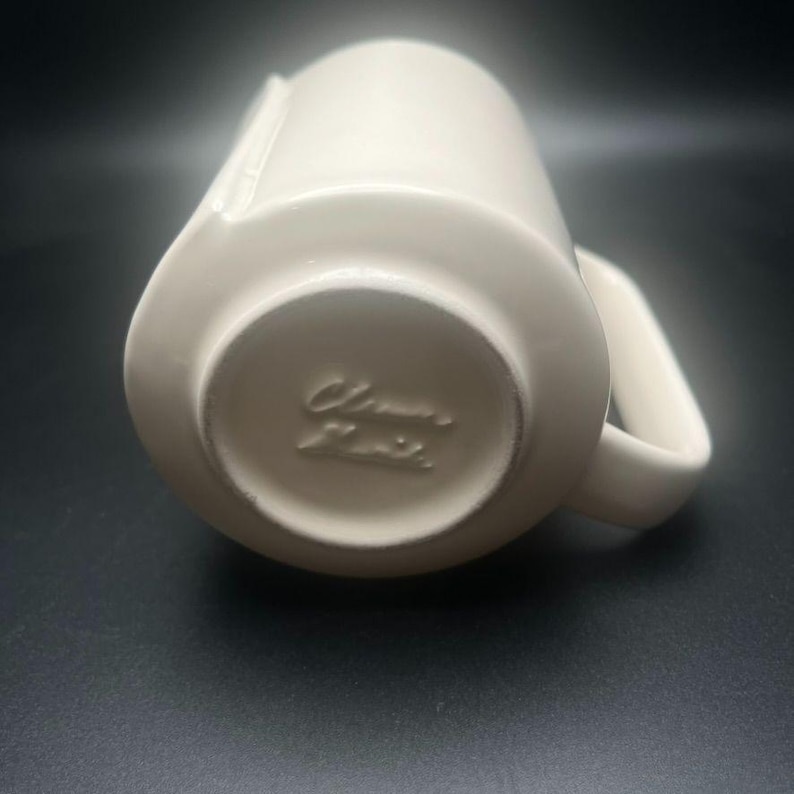 May include: Cream-colored ceramic mug with a handle and a spout. The mug has a rounded base and a cylindrical body. The bottom of the mug has the embossed text "Clamart Paris". The mug is set against a dark background.