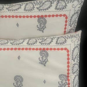 May include: Two white pillowcases with a repeating gray paisley and floral pattern. A red beaded border frames the center design. The pillowcases are a decorative bedding item.