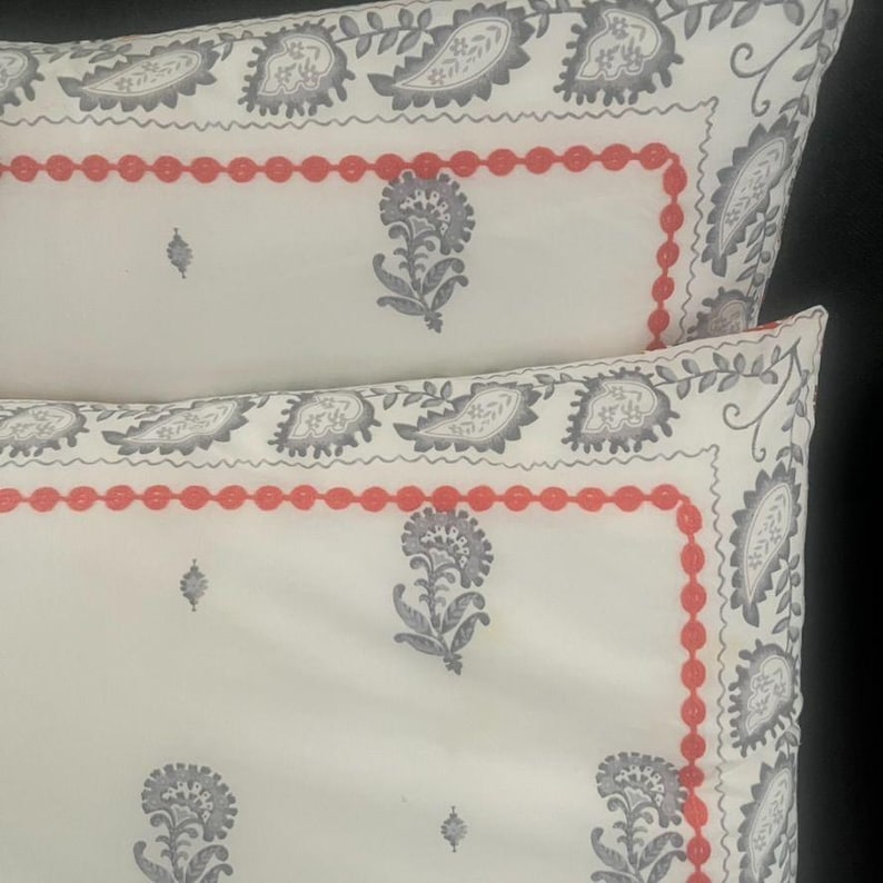 May include: Two white pillowcases with a repeating gray paisley and floral pattern. A red beaded border frames the center design. The pillowcases are a decorative bedding item.