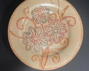 May include: A beige ceramic plate with a floral design. The flowers are made of red and black dots, with orange stems and swirls. The rim is decorated with small orange dots.