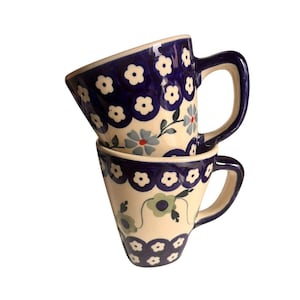 May include: Two ceramic mugs stacked on top of each other. The mugs are cream-colored with a blue floral pattern and blue handles. The floral pattern includes white and blue flowers with green leaves.