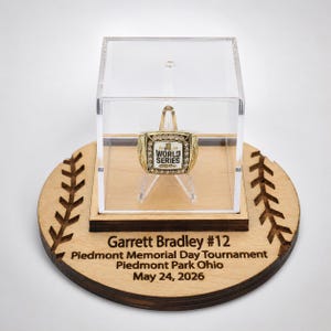 May include: A baseball World Series ring displayed in a clear acrylic case, set on a wooden base. The ring is gold-toned with a white center and the words "WORLD SERIES". The base is engraved with "Garrett Bradley #12" and tournament details.