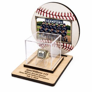May include: A wooden display stand featuring a circular baseball-themed backboard with a team photo labeled 2026 10U TOURNAMENT CHAMPIONS. A clear acrylic cube houses a gold championship ring. The base is engraved with Jared Williams #33, Willow Brooke Braves, Pinto World Series Champions, July 3, 2026.
