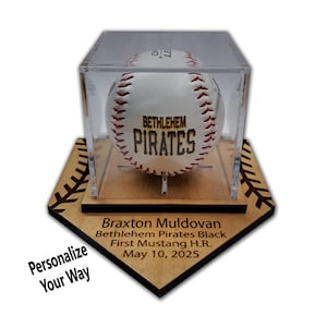 May include: A baseball in a clear acrylic display case, resting on a wooden base. The baseball is white with red stitching and the words "BETHLEHEM PIRATES" in brown. The base has engraved text: "Braxton Muldovan, Bethlehem Pirates Black, First Mustang H.R., May 10, 2025."