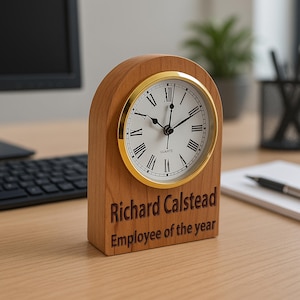 May include: A wooden desk clock with a gold rimmed face and Roman numeral markings. The clock is engraved with the text "Richard Calstead Employee of the year".