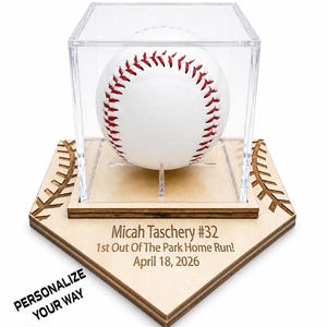 May include: A baseball in a clear acrylic display case, resting on a wooden home plate-shaped base. The baseball is white with red stitching. The base has engraved text: "Micah Taschery #32 1st Out Of The Park Home Run! April 18, 2026".