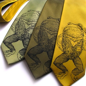 May include: Three silk neckties in shades of green and gold, each featuring a detailed black illustration of a frog in a waistcoat and trousers. The ties vary in color from light olive to mustard yellow.