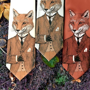 May include: Three neckties with a fox illustration wearing a suit. The ties are in shades of cream, brown, and rust. The fox is depicted with a detailed face and is wearing a suit with a pocket square. The ties are arranged on a bed of purple foliage.