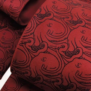 May include: Close-up of a fabric with a repeating pattern of black octopus illustrations on a deep red background. The fabric is folded, showing the pattern on multiple surfaces. The design is detailed and artistic.