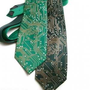 以下が含まれることがあります： 回路基板のデザインのネクタイ2本。1本はエメラルドグリーンに金色の回路基板模様、もう1本は濃いフォレストグリーンに金色の回路基板模様。ネクタイは白い背景に表示されています。