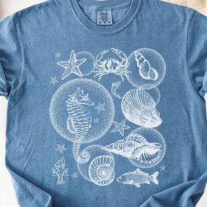 May include: Blue jean-colored t-shirt featuring a white illustration of various sea creatures, including a seahorse, crab, starfish, and seashells. The design is printed in a circular pattern. The shirt is made of soft material.
