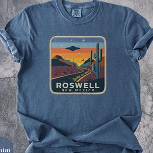 May include: A denim blue short-sleeve t-shirt featuring a graphic print of a desert landscape with a saguaro cactus, a winding road, and a flying saucer. The text reads "Stay Wild, Stay Weird," "ROSWELL," and "NEW MEXICO" in a retro-style design.