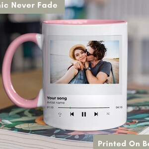 May include: White ceramic mug with a pink interior and handle. The mug features a photo of a couple kissing, with a music player interface below. Text on the mug reads "Your song" and "Printed On Both Sides". The text "Graphic Never Fade" is also visible.