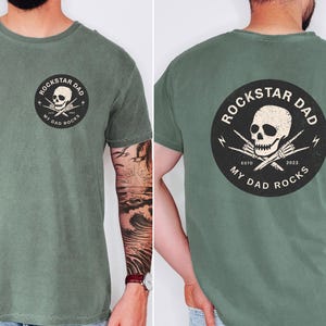 May include: Moss green t-shirt with a circular graphic featuring a skull, crossed drumsticks, and the text "ROCKSTAR DAD MY DAD ROCKS ESTD 2023". The design is on the front and back of the shirt.