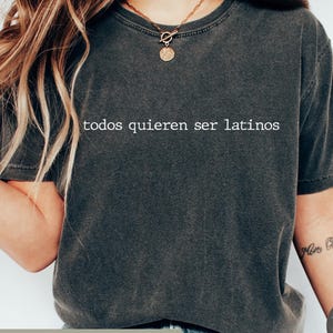 May include: Dark gray t-shirt with the text "todos quieren ser latinos" in white. The shirt has short sleeves and a crew neck. The image also shows a bracelet and a tattoo on the arm.