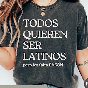 May include: Dark gray t-shirt with the text "TODOS QUIEREN SER LATINOS pero les falta SAZ&Oacute;N" in white. The shirt has a vintage, worn look. The words are stacked vertically. The shirt is part of a clothing collection.