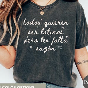 May include: Dark gray t-shirt with the text "todos quieren ser latinos pero les falta saz&oacute;n" in white script. The shirt has a vintage, worn look and is a casual style.