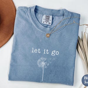 May include: Blue jean-colored t-shirt with the words "let it go" and a dandelion graphic. A gold necklace with a pendant rests on the shirt. A brown hat and dried plant are in the background.