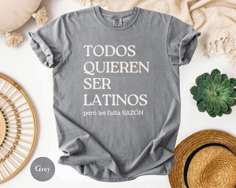 May include: A gray t-shirt with the text "TODOS QUIEREN SER LATINOS pero les falta SAZ&Oacute;N" in white. The shirt is displayed on a white surface with a straw hat, a succulent plant, and decorative items.
