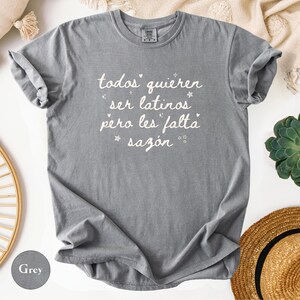 May include: A grey t-shirt with the text "todos quieren ser latinos pero les falta saz&oacute;n" in white script. The shirt is folded, with rolled-up sleeves. The shirt is on a white surface with a straw hat, a succulent plant, and a beige blanket.