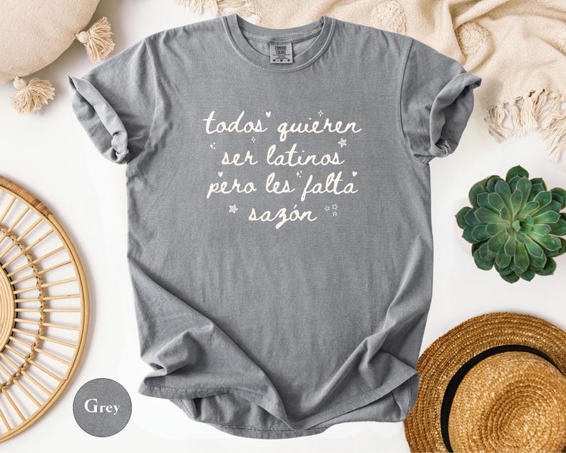 May include: A grey t-shirt with the text "todos quieren ser latinos pero les falta saz&oacute;n" in white script. The shirt is folded, with rolled-up sleeves. The shirt is on a white surface with a straw hat, a succulent plant, and a beige blanket.