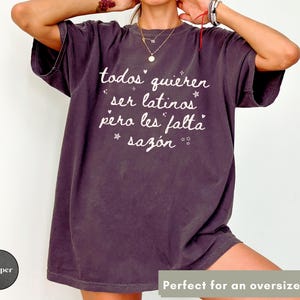 May include: A dark purple oversized t-shirt with white text that reads "todos quieren ser latinos pero les falta saz&oacute;n". The shirt has a vintage, worn look. The text is in a handwritten style with star accents.