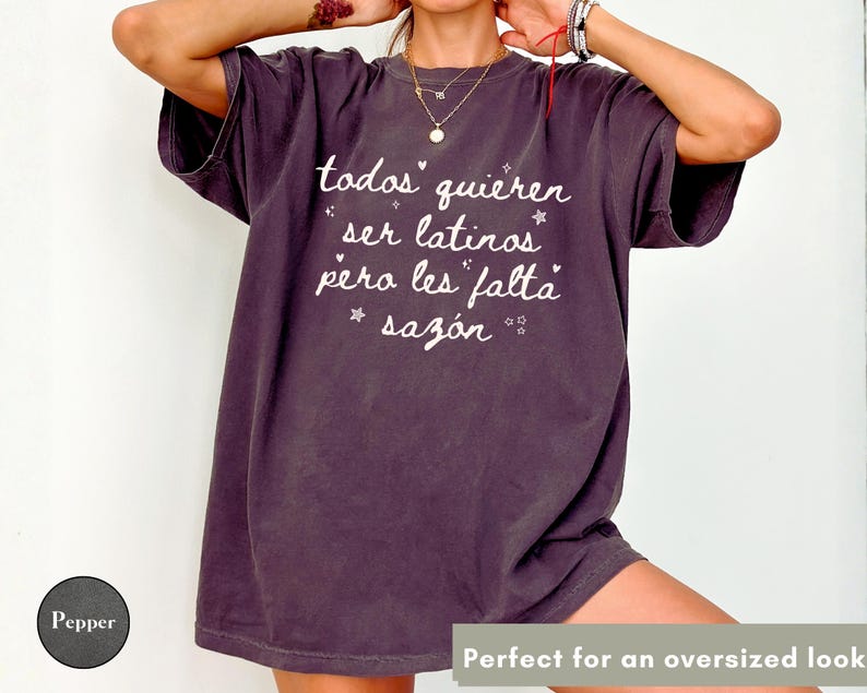 May include: A dark purple oversized t-shirt with white text that reads "todos quieren ser latinos pero les falta saz&oacute;n". The shirt has a vintage, worn look. The text is in a handwritten style with star accents.