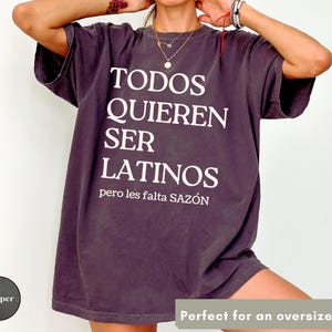 May include: Dark purple oversized t-shirt with white text that reads "TODOS QUIEREN SER LATINOS pero les falta SAZ&Oacute;N". The shirt has short sleeves and a relaxed fit.