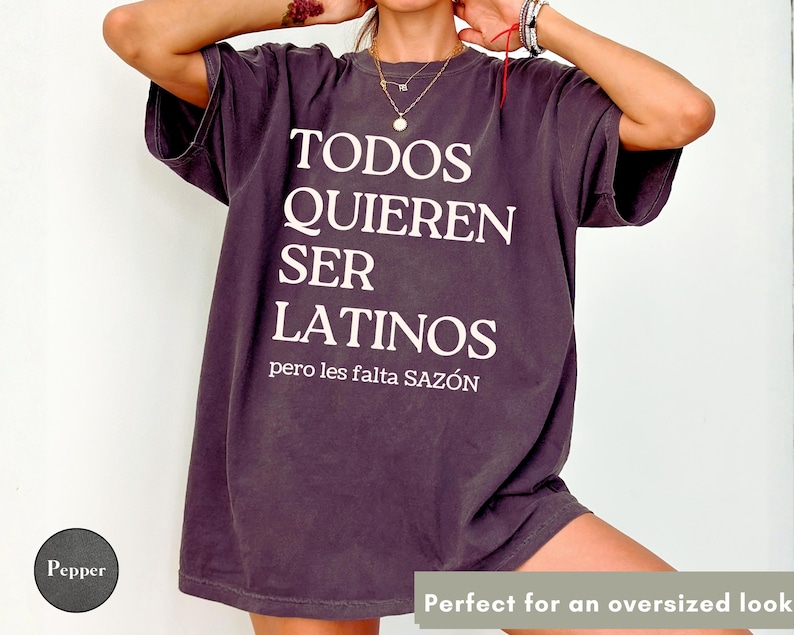 May include: Dark purple oversized t-shirt with white text that reads "TODOS QUIEREN SER LATINOS pero les falta SAZ&Oacute;N". The shirt has short sleeves and a relaxed fit.