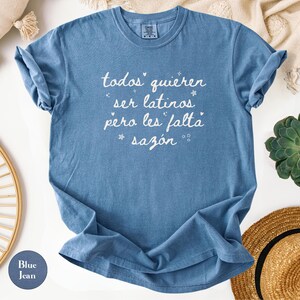 May include: Blue jean-colored t-shirt with white script text that reads "todos quieren ser latinos pero les falta saz&oacute;n." The shirt is folded, with rolled-up sleeves. The shirt is surrounded by a straw hat, a succulent plant, and a decorative pillow.