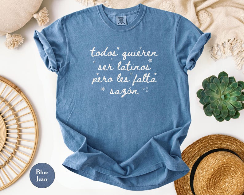 May include: Blue jean-colored t-shirt with white script text that reads "todos quieren ser latinos pero les falta saz&oacute;n." The shirt is folded, with rolled-up sleeves. The shirt is surrounded by a straw hat, a succulent plant, and a decorative pillow.
