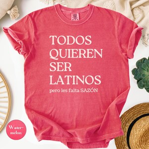 May include: A watermelon-colored t-shirt with the text "TODOS QUIEREN SER LATINOS pero les falta SAZ&Oacute;N" in white. The shirt is folded, with rolled-up sleeves. A small circle with the word "Watermelon" is in the lower left corner.