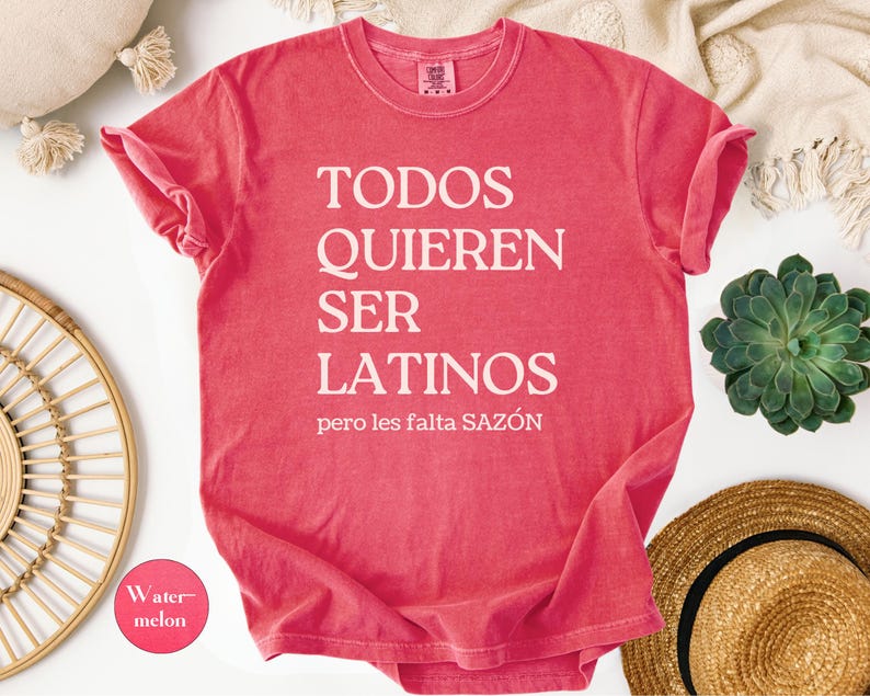May include: A watermelon-colored t-shirt with the text "TODOS QUIEREN SER LATINOS pero les falta SAZ&Oacute;N" in white. The shirt is folded, with rolled-up sleeves. A small circle with the word "Watermelon" is in the lower left corner.