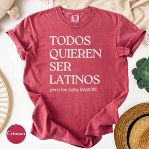 May include: Crimson-colored t-shirt with the text "TODOS QUIEREN SER LATINOS pero les falta SAZ&Oacute;N" in white. The shirt is displayed on a white surface with decorative items, including a straw hat and a succulent plant.