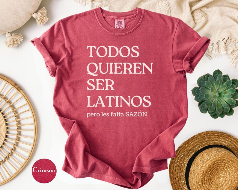 May include: Crimson-colored t-shirt with the text "TODOS QUIEREN SER LATINOS pero les falta SAZ&Oacute;N" in white. The shirt is displayed on a white surface with decorative items, including a straw hat and a succulent plant.