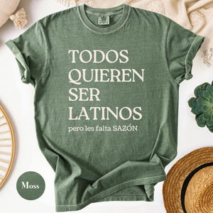 May include: A moss green t-shirt with the text "TODOS QUIEREN SER LATINOS pero les falta SAZ&Oacute;N" in white. The shirt is folded, with a round neck and short sleeves. A round moss green color swatch is in the lower left corner.