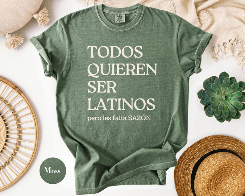 May include: A moss green t-shirt with the text "TODOS QUIEREN SER LATINOS pero les falta SAZ&Oacute;N" in white. The shirt is folded, with a round neck and short sleeves. A round moss green color swatch is in the lower left corner.