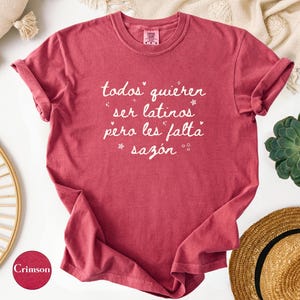May include: Crimson-colored t-shirt with rolled-up sleeves. The shirt features white script that reads "todos quieren ser latinos pero les falta saz&oacute;n." The shirt is displayed on a white surface with decorative items.