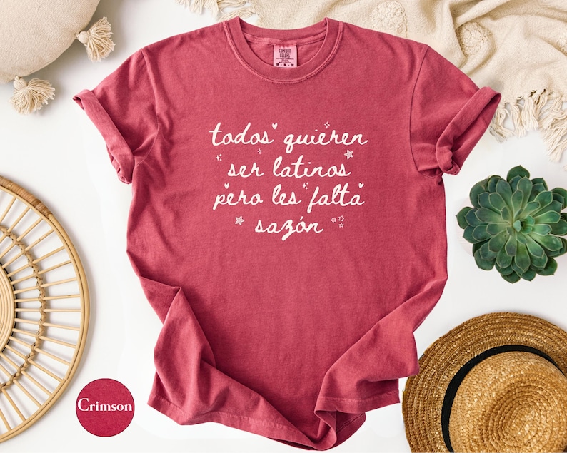 May include: Crimson-colored t-shirt with rolled-up sleeves. The shirt features white script that reads "todos quieren ser latinos pero les falta saz&oacute;n." The shirt is displayed on a white surface with decorative items.