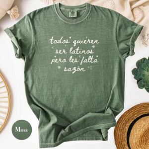 May include: A moss green t-shirt with rolled-up sleeves. The shirt features white text that reads "todos quieren ser latinos pero les falta saz&oacute;n" with star accents. The shirt is part of a clothing collection.