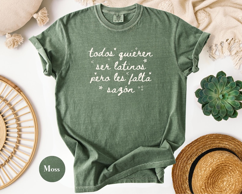 May include: A moss green t-shirt with rolled-up sleeves. The shirt features white text that reads "todos quieren ser latinos pero les falta saz&oacute;n" with star accents. The shirt is part of a clothing collection.