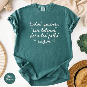 May include: Blue spruce t-shirt with the text "todos quieren ser latinos pero les falta saz&oacute;n" in white script. The shirt is folded, with a round tag that says "Blue Spruce". The shirt is surrounded by decorative items.