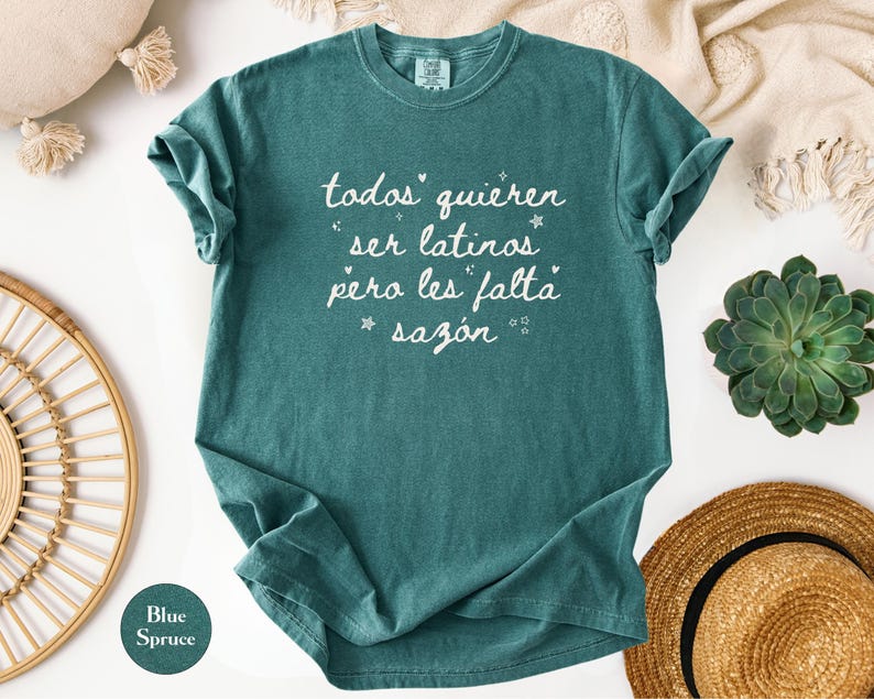 May include: Blue spruce t-shirt with the text "todos quieren ser latinos pero les falta saz&oacute;n" in white script. The shirt is folded, with a round tag that says "Blue Spruce". The shirt is surrounded by decorative items.