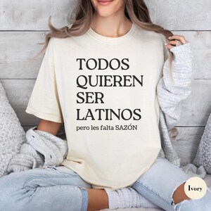 May include: Ivory-colored t-shirt with the text "TODOS QUIEREN SER LATINOS pero les falta SAZ&Oacute;N" in black. The shirt is a relaxed fit, with a crew neck. The model is wearing light-wash jeans.